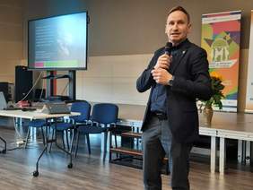 Pokaż zdjęcie: Mężczyzna w garniturze przemawia do mikrofonu. Za nim wisi tablica multimedialna. 