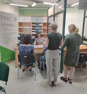 Pokaż zdjęcie: Przedstawicielka PFRON prowadzi rozmowę z dwiema osobami siedzącymi przy biurku. Obok kolejni uczestnicy czekają w kolejce po konsultacje. W tle widoczny roll-up informacyjny i regały z dokumentami w urzędowym wnętrzu.