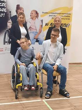 Pokaż zdjęcie: Na tle banera reklamującego kampanię społeczną "W kontakcie" znajdują się cztery osoby: dwoje dzieci i dwie kobiety. Dziewczynka siedzi na wózku inwalidzkim, a obok niej na krześle siedzi chłopiec. Panie stoją za dziećmi. 