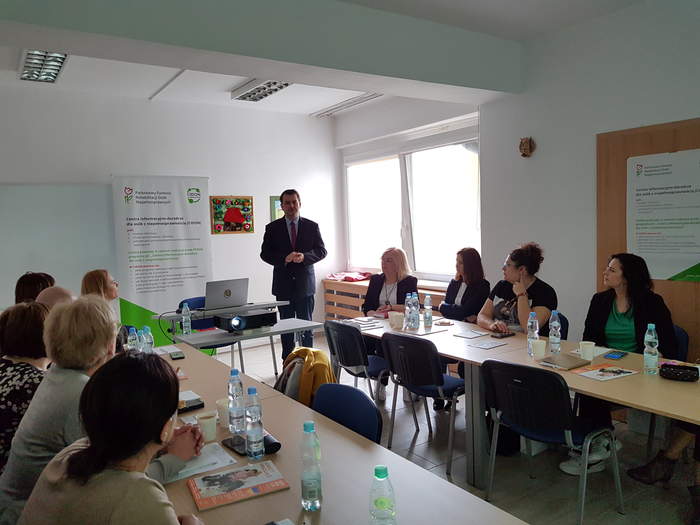 Pokaż zdjęcie: Na sali przy stołach siedzą pracownicy OPS, na środku stoi Dyrektor i przemawia, uczestnicy słuchają, w tle jest okno, widok na miasto