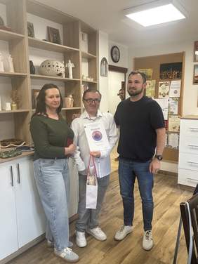 Pokaż zdjęcie: Trzy osoby pozują do wspólnej fotografii w jasnym wnętrzu z drewnianymi półkami i dekoracjami. Osoba stojąca w centrum trzyma dyplom i torbę z upominkiem. W tle znajdują się półki z ozdobami, zegar ścienny oraz tablica z materiałami informacyjnymi. Zdjęcie dokumentuje moment wręczenia wyróżnienia lub podziękowania.