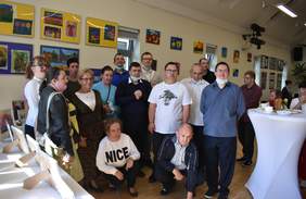 Pokaż zdjęcie: Grupa uśmiechniętych osób stoi na tle  prac plastycznych wiszących na ścianie