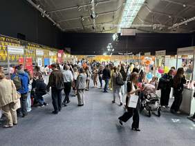 Pokaż zdjęcie: Targi pracy w hali sportowej z licznymi stoiskami firm i tłumem odwiedzających; widoczne kolorowe balony, wózki dziecięce i szyldy firm m.in. Adecco, Auchan Polska i Avon Cosmetic Polska.