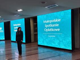 Pokaż zdjęcie: Kobieta stojąca w holu budynku na tle dużych ekranów z napisem „Małopolskie Spotkanie Opłatkowe, 16 grudnia 2025”, prezentujących świąteczną grafikę wydarzenia.