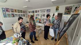 Pokaż zdjęcie: Grupa osób stojąca w pracowni artystycznej obok dużej czarno-białej fotografii ustawionej na sztaludze. Jedna z osób trzyma dyplom i torbę z nagrodą. W tle widoczne są kolorowe prace plastyczne i materiały artystyczne.