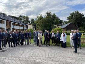Pokaż zdjęcie: Na zewnątrz, na tle nowego budynku stoją zaproszeni goście. Wszyscy ubrani na galowo. Po środku ubrany w granatowy garnitur, białą koszulę i krawat dyrektor. Ksiądz stojący po prawej stronie modli się. Jest słoneczna pogoda.