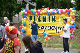 Pokaż zdjęcie: Na skraju boiska do gry w koszykówkę ustawiony został baner w kolorze żółtym z napisem: „Piknik integracyjny” oraz logotypem Warsztatów Terapii Zajęciowej w Szamotułach. Na obrzeżach baneru przymocowane zostały duża liczba różnokolorowych balonów. Na boisku stoi grupa osób, które dotarły na piknik integracyjny. Wśród tej grupy możemy dostrzec także klauna.
