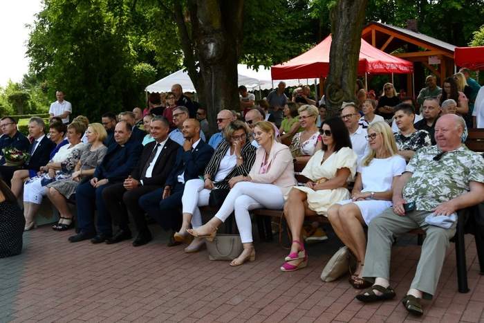 Pokaż zdjęcie: Grupa kilkudziesięciu osób siedzi na drewnianych ławkach oraz krzesłach ustawionych w kilku rzędach pod wysokimi drzewami. Z tyłu ustawione są trzy namioty, w pobliżu których zgromadziła się duża grupa osób.