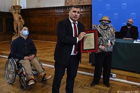 Pokaż zdjęcie: Mężczyzna w granatowym garniturze , białej koszuli i ciemnym krawacie trzyma w ręku  drewnianą tablicę z aluminiową blaszką z grawerem. Za nim siedzi osoba na wózku inwalidzkim.