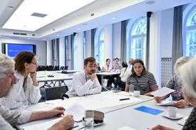 Pokaż zdjęcie: Ludzie siedzą przy kilku stołach, dyskutując i czytając dokumenty. W pomieszczeniu panuje atmosfera skupienia i współpracy.