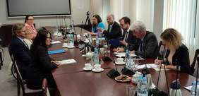 Pokaż zdjęcie: W sali konferencyjnej przy dużym stole siedzi 9 osób. Trwa dyskusja. Przed rozmawiającymi leżą dokumenty. W tle ekran.