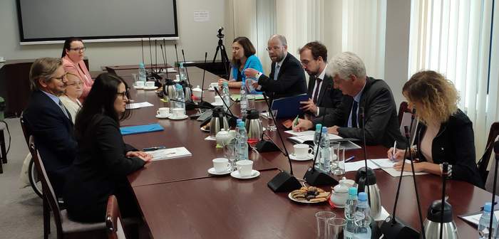 Pokaż zdjęcie: W sali konferencyjnej przy dużym stole siedzi 9 osób. Trwa dyskusja. Przed rozmawiającymi leżą dokumenty. W tle ekran.