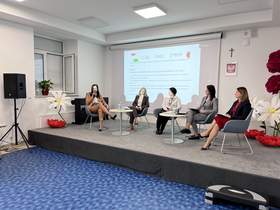 Sala konferencyjna w jasnych popielatych kolorach, podłoga niebieska, 5 kobiet siedzi na scenie, rozmawiają, za nimi duży ekran z wyświetloną treścią