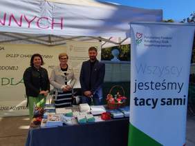 Pokaż zdjęcie: Trzy osoby stoją przy stoisku informacyjnym PFRON podczas wydarzenia plenerowego. Na stole widoczne są broszury, ulotki i materiały promujące wsparcie dla osób z niepełnosprawnościami. Obok znajduje się baner z hasłem „Wszyscy jesteśmy tacy sami”.