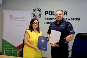 Pokaż zdjęcie: Dwie osoby prezentują podpisane dokumenty w siedzibie Komendy Wojewódzkiej Policji w Lublinie. W tle widoczne są logotypy Policji i PFRON oraz roll-up funduszu.