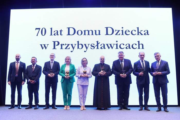 Pokaż zdjęcie: Na scenie w rzędzie stoi 9 osób, pośrodku dwie kobiety, ksiądz i sześciu mężczyzn. Osoby te trzymają w ręku pamiątkowe grawertony. W tle prezentacja z hasłem, 70-lecie Domu Dziecka w Przybysławicach.