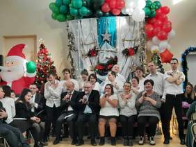 Pokaż zdjęcie: Na scenie znajdują się osoby z niepełnosprawnościami, które prezentują zebranym występ artystyczny. Niektórzy śpiewają, a inni gestykulują. W tle widać świąteczne dekoracje.  