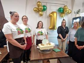 Pokaż zdjęcie: Grupa uśmiechniętych osób stoi wokół stolika. Na stoliku ustawiony jest tort ze świeczką i dekoracją w kształcie filiżanki. Na ścianie jest dekoracja z balonów. 