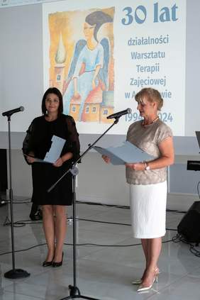 Pokaż zdjęcie: Dwie kobiety stojące przy mikrofonach podczas uroczystości. Trzymają w rękach teczki z dokumentami i przemawiają do publiczności. W tle, na dużym ekranie, wyświetlany jest plakat z napisem „30 lat działalności Warsztatu Terapii Zajęciowej w Augustowie 1994-2024” oraz grafiką przedstawiającą anioła siedzącego na dachu domów. Kobieta po lewej stronie ma na sobie czarną sukienkę, a kobieta po prawej jasną bluzkę i białą spódnicę.