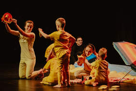 Pokaż zdjęcie: Fragment przedstawienia, na scenie cztery mamy wraz z dziećmi bawiące się na plaży. Wszyscy ubrani są w żółte stroje.