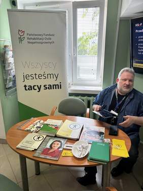 Pokaż zdjęcie: Mężczyzna siedzi przy okrągłym stoliku, na którym znajdują się broszury, teczki i materiały informacyjne. Obok stoi baner z napisem „Wszyscy jesteśmy tacy sami”.)