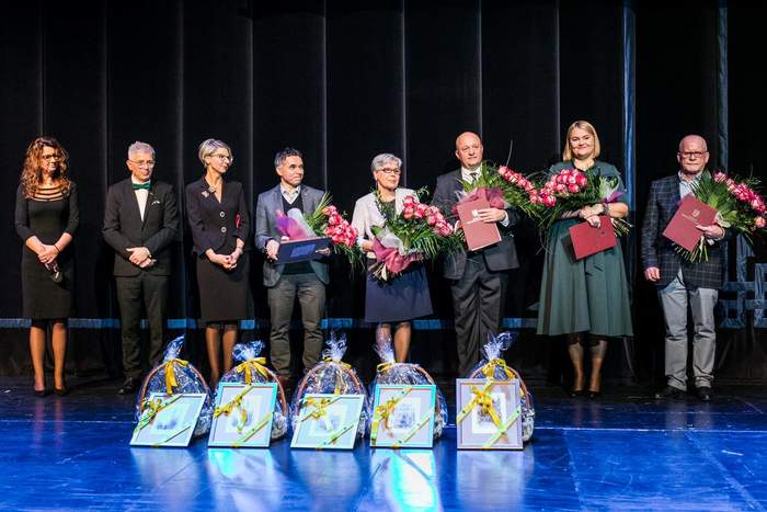 Pokaż zdjęcie: Na scenie teatralnej stoi czterech mężczyzn i 4 kobiety. Pięcioro z nich trzyma duże bukiety róż i listy gratulacyjne. Wszyscy się uśmiechają. Są ubrani na są galowo. Przed nimi leżą nagrody w postaci wypełnionych koszy wiklinowych, o które