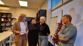 Pokaż zdjęcie: Na zdjęciu cztery osoby, trzy kobiety i mężczyzna. Mężczyzna ubrany w jeansową koszulę, trzyma ręce skrzyżowane na piersi. Kobiety żywo ze sobą rozmawiają.