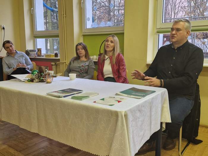 Pokaż zdjęcie: Przy stole z białym obrusem na którem leża ulotki siedzą trzy kobiety i jeden mężczyzna. 