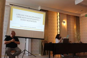 Pokaż zdjęcie: W sali konferencyjnym po lewej stronie siedzi na krześle tłumacz języka migowego, po prawej za stołem prezydialnym siedzi kobieta z mikrofonem, w tle po lewej na ekranie wyświetlana jest prezentacja