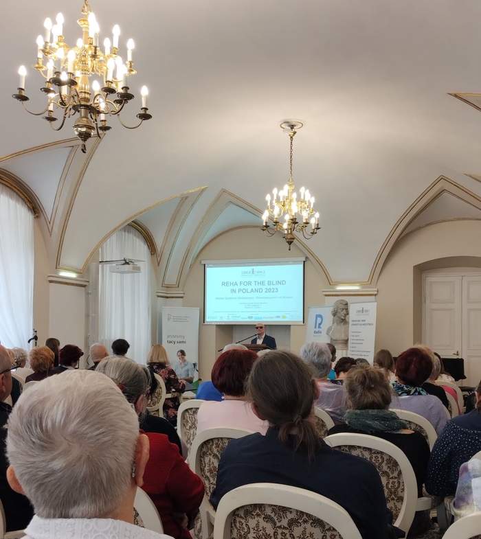 Pokaż zdjęcie: Grupa kilkunastu osób siedzi w sali z zaokrąglonym sufitem i dwoma podwieszanymi żyrandolami. Na mównicy stoi mężczyzna w ciemnych okularach, który przemawia do mikrofonu. W tle widoczne są banery Fundacji Szansa dla Niewidomych, Radia Poznań oraz Państwowego Funduszu Rehabilitacji Osób Niepełnosprawnych.