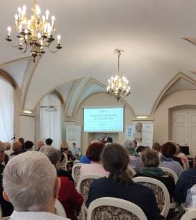 Pokaż zdjęcie: Grupa kilkunastu osób siedzi w sali z zaokrąglonym sufitem i dwoma podwieszanymi żyrandolami. Na mównicy stoi mężczyzna w ciemnych okularach, który przemawia do mikrofonu. W tle widoczne są banery Fundacji Szansa dla Niewidomych, Radia Poznań oraz Państwowego Funduszu Rehabilitacji Osób Niepełnosprawnych.