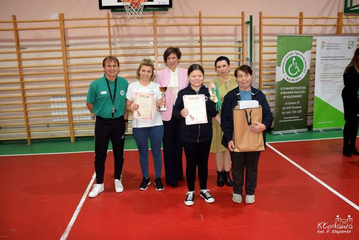 Pokaż zdjęcie: W hali sportowej, obok banerów: Stowarzyszenia „Sprawni Inaczej” oraz Centrum Informacyjno-Doradczego dla Osób z Niepełnosprawnością, stoi pięć kobiet oraz jeden mężczyzna. Wśród tych osób są zawodnicy z dyplomami, pucharem i nagrodami rzeczowymi.