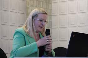 Pokaż zdjęcie: Kobieta blondynka w zielonej marynarce trzyma mikrofon i spogląda w ekran laptopa.