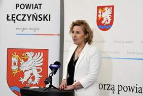 Pokaż zdjęcie: Kobieta w białej marynarce stoi przed mównicą. Za nią biała ściana, na której herb na czerwonym tle biały gryf, pod nim niebieska wstęga 