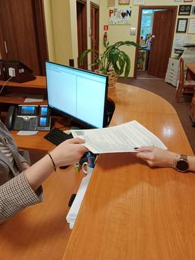 Pokaż zdjęcie: Dwie osoby podają sobie wniosek o dofinansowanie. Widać jedynie dłonie z dokumentem. W tle jest także lada, komputer, kwiat i inne urządzenia biurowe. 