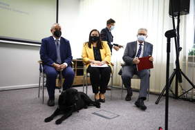 Pokaż zdjęcie: Grupa 3 osób, siedzących na krzesłach w sali konferencyjnej. Jednej z nich towarzyszy pies przewodnik. 