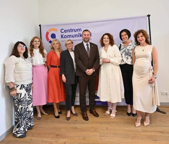Pokaż zdjęcie: Grupa ośmiu osób stoi w pomieszczeniu przed banerem „Centrum Komunikacji”; formalny strój, jasne ściany i drewniana podłoga, ujęcie pozowane.