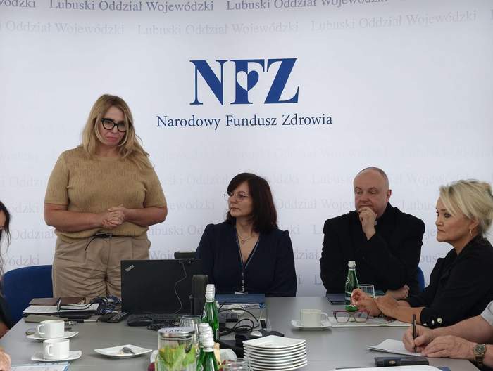 Pokaż zdjęcie: Na tle logo NFZ stoi kobieta i przemawia, a obok niej przy stole siedzą uczestnicy kapituły