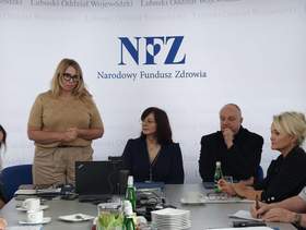 Na tle logo NFZ stoi kobieta i przemawia, a obok niej przy stole siedzą uczestnicy kapituły