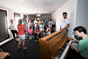 Pokaż zdjęcie: Grupa uczestników oczekuje na warsztaty muzyczne, poprzedzające koncerty konkursowe, na pierwszym planie jeden z uczestników przygrywa na pianinie