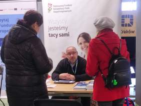 Pokaż zdjęcie: Dwie kobiety stoją przy stoisku informacyjnym PFRON. Mężczyzna siedzący przy stoliku udziela informacji. Za stoiskiem rollup z logo PFRON.