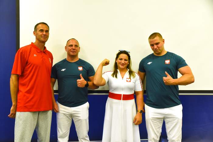 Pokaż zdjęcie: Dwóch mężczyzn w białych spodniach i zielonych koszulkach, jeden mężczyzna w czerwonej koszulce i jasnych spodniach, kobieta w białej sukience