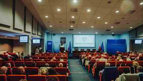 Pokaż zdjęcie: Wypełniona ludźmi sala konferencyjna z rzędami czerwonych foteli skierowanych w stronę sceny. Na podium widoczna jest mównica, duży jasny ekran, niebieskie panele informacyjne oraz flagi, w tym flaga Unii Europejskiej.