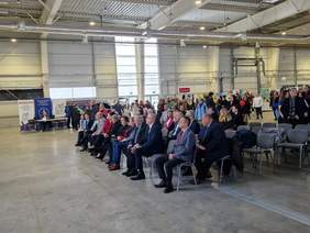 Pokaż zdjęcie: Zdjęcie hali targowej na krzesełkach siedzą zaproszeni goście, trwa otwarcie targów, część oficjalna.