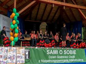 Pokaż zdjęcie: scena w plenerze na której siedzą osbu uczestniczące w przeglądzie artystycznym. Panie mają piękne długie społdnice  w stylu hiszpańskim, a panowie w pomarańczowych koszulach pod krawatem siedzą na scenie i śpiewają