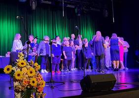 Pokaż zdjęcie: Grupa osób stoi na scenie oświetlonej zielonym światłem, przygotowanej do występu. Na przodzie widoczny bukiet słoneczników i róż. W tle mikrofony i kurtyna sceniczna, a ludzie rozmawiają i ustawiają się w grupie.  
