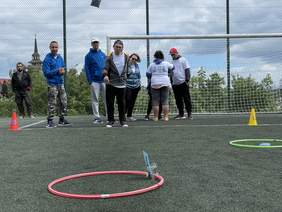Pokaż zdjęcie: Mężczyzna ubrany w trampki ciemne spodnie dresowe bluzę oraz czapkę założoną tył na przód wyrzuca sakiewkę do leżącego przed nim kółka z napisem 4. Za nim stoją inni zawodnicy. W tle bramka ta piłkarska i siatka otaczająca boisko.