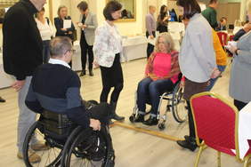 Pokaż zdjęcie: Zdjęcie sali w której odbywają się warsztaty tematyczne, zdrowe oso by próbuja przejechac na wózku mały próg wysokości 3 cm. Z tyłu asekurują je osoby, wokoło stoją ludzie i się przyglądają