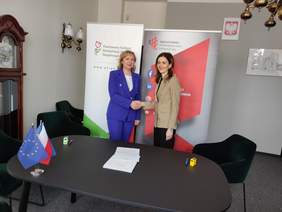 Pokaż zdjęcie: Na tle dwóch banerów z logo PFRON oraz WSPiA w Lublinie stoją dwie kobiety wymieniające uścisk dłoni. Zdjęcie w sali konferencyjnej