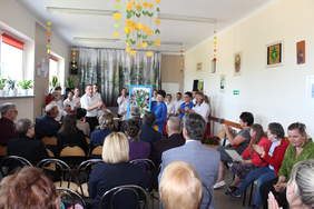 Pokaż zdjęcie: Na środku kobieta ubrana na niebiesko stoi i trzyma obraz z przyklejonymi imionami dzieci, z tyłu ubrana na biało grupa wychowanków ośrodka